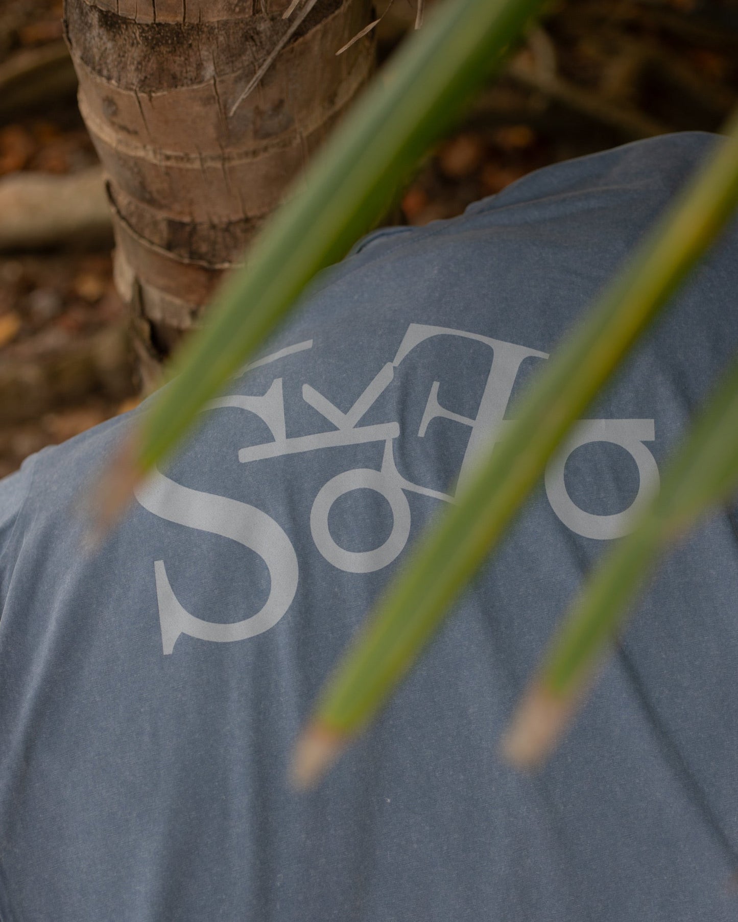 blue shirt with a logo, surrounded by green palm tree leaves and blurred natural background